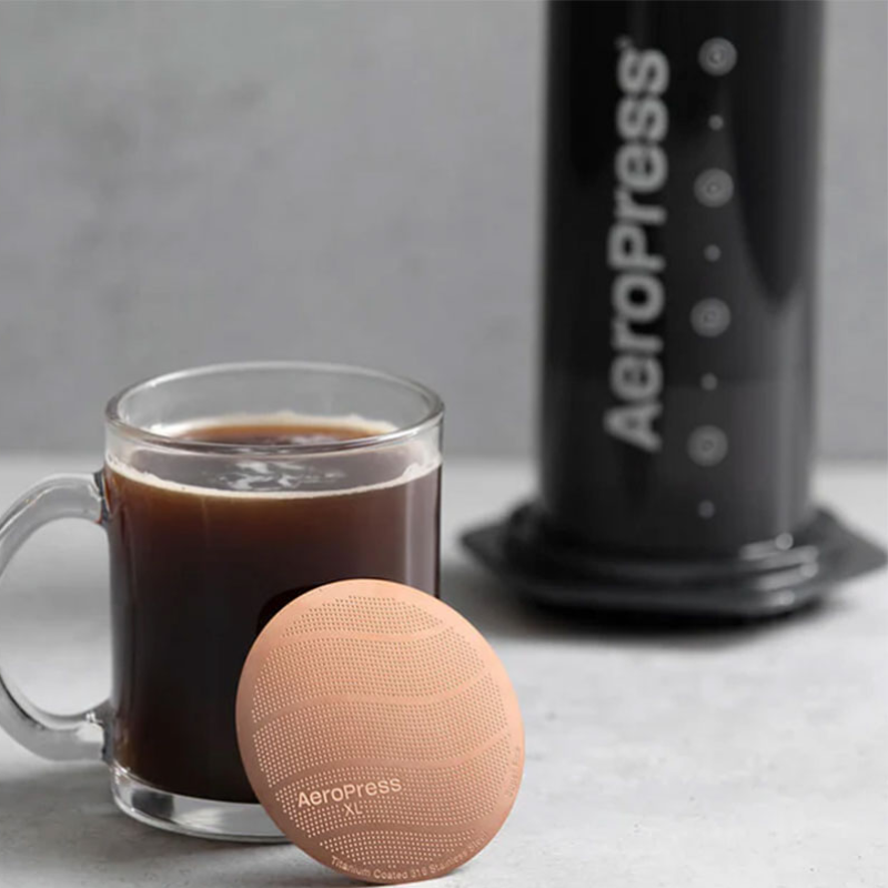 AeroPress Gold Tone Metalfilter XL — XL-version af det kobberfarvede metalfilter placeret ved en kop kaffe med AeroPress i baggrunden, tæt beskåret produktfoto.