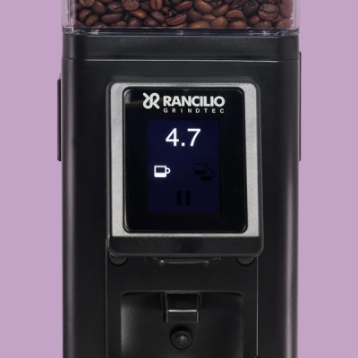 Rancilio kaffekværn med digitalt display og kværnede bønner i beholderen, set forfra mod en lilla baggrund.